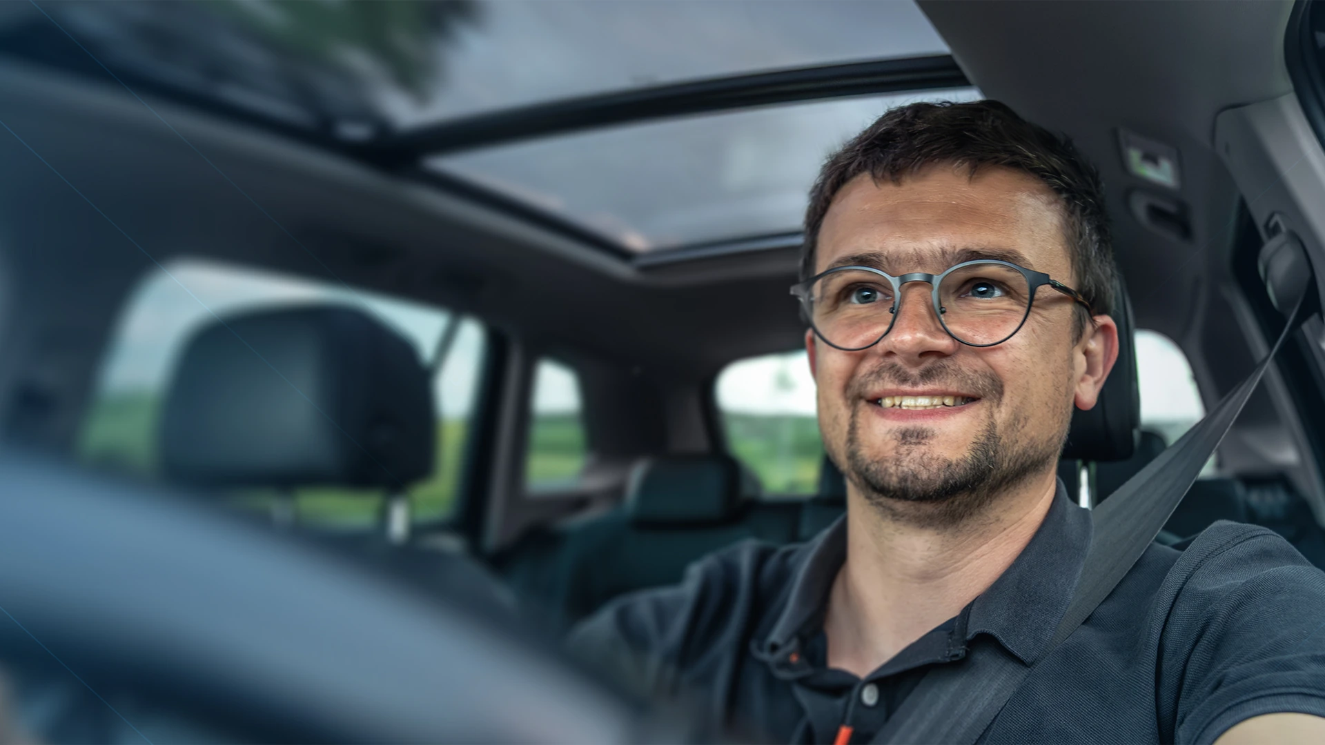 Homem sorridente de óculos dirigindo um carro com teto solar, paisagem verde através das janelas.