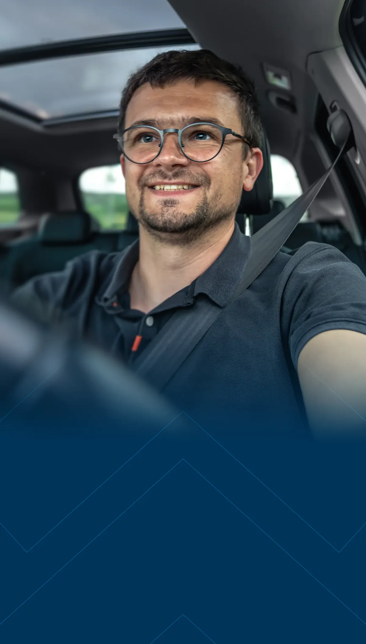 Homem sorridente de óculos dirigindo um carro com teto solar, paisagem verde através das janelas.